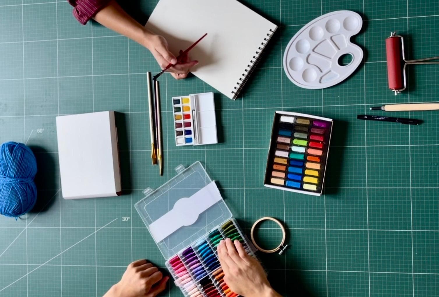 Matériel d'atelier vu de dessus : aquarelle, pastels, fils de broderie, pinceaux